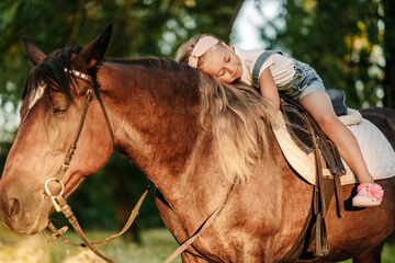 A little blonde girl with long hair lies and hugs astride a horse in the park at sunset in autumn. Autumn horse ride. Friendship of a girl and a horse.