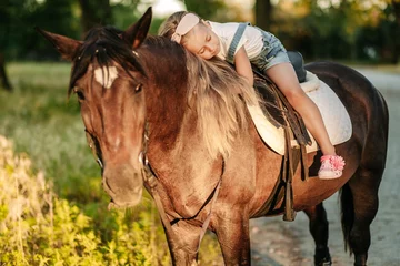 Foto op Canvas A little blonde girl with long hair lies and hugs astride a horse in the park at sunset in autumn. Autumn horse ride. Friendship of a girl and a horse. © sergo321