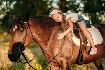 Fototapeta premium A little blonde girl with long hair lies and hugs astride a horse in the park at sunset in autumn. Autumn horse ride. Friendship of a girl and a horse.