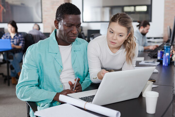 Attractive friendly girl helping African male colleague in work in modern coworking space