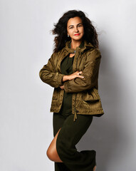 Smiling mature curly brunette woman model in stylish overall and jacket stands at the wall holding arms crossed at chest