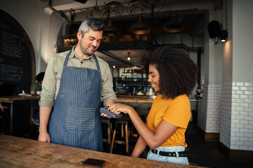 Beautiful ethnic woman tapping card making payment for coffee to handsome waiter in trendy cafe.