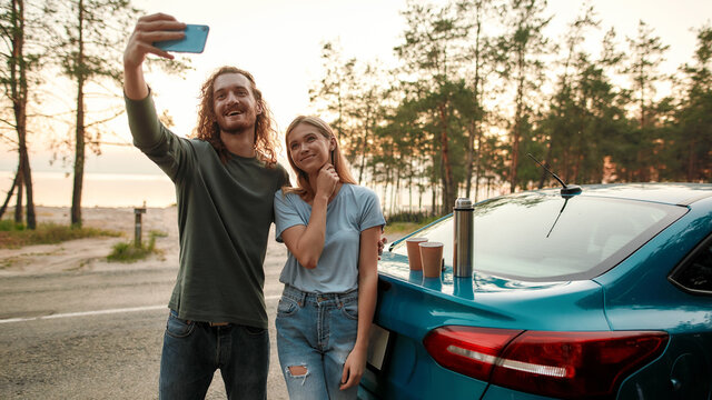 Cheerful man using smartphone while taking selfie together with attractive young woman after repairing broken car on the road side