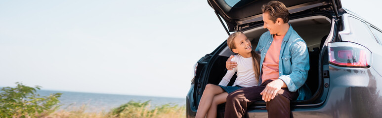 Panoramic shot of father hugging daughter in car trunk near seaside