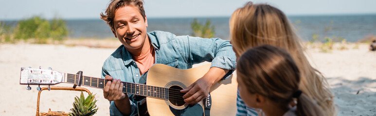 Horizontal image of man playing acoustic guitar near family on beach
