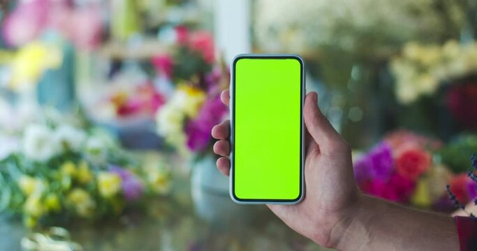 Male hand in modern flower store, shop showing chroma key. Man florist holding smartphone with green screen. Blurred blossom plant background. Busines, entreprenuership concept.
