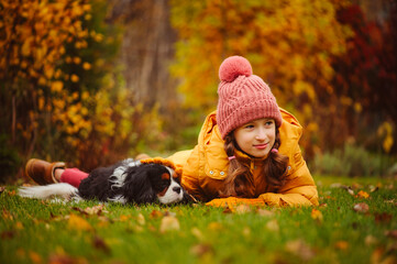 Happy kid girl walking and playing with her cavalier king charles spaniel dog in autumn garden or...