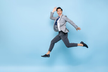 Fototapeta na wymiar Full length fun portrait of happy energetic young Asian businessman jumping in mid-air isolated on studio blue background with copy space