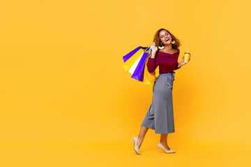 Full length portrait of smiling young African American woman holding shopping bags and drink cup walking in isolated studio yellow background with copy space
