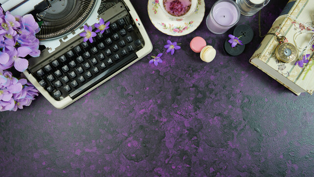 Purple theme vintage writers desktop workspace with typewriter, lavendar tea, hibiscus flowers and old books on stylish purple textured background. Top view blog hero header creative flat lay.