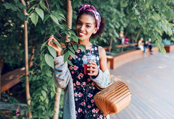 Smiling black woman in stylish hipster clothes grey cardigan walking in sunny park and  enjoying   weekends, Trendy bamboo bag .  Drinking lemonade.