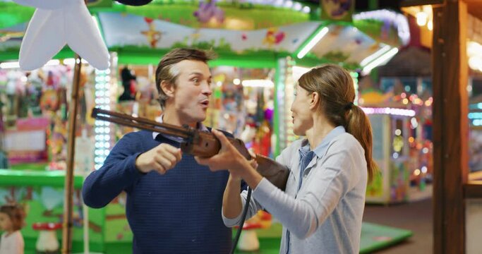 Authentic shot of a happy carefree smiling couple in love is having fun to play fair shooting games and win in amusement park with luna park lights at night.