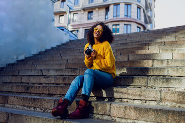 Fototapeta premium Stylish inspired American female student sitting on stairs and looking forward. . Holding retro film camera. Urban background. Autumn trends.