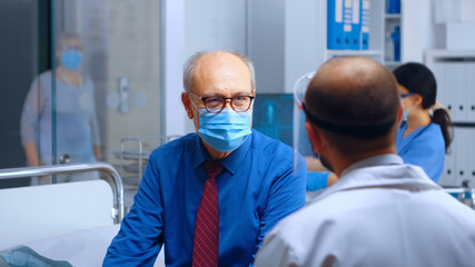 Fototapeta na wymiar Physician talking with patient in mask during COVID 19 health crisis. Medical healthcare system global pandemic. Medicine worker offering consultation to elderly retired old man. Handheld slow motion