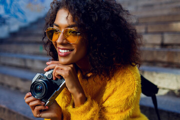 Stylish inspired African woman sitting on stairs and dreaming. Holding retro film camera. Urban...