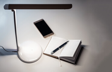 A smartphone and open diary with a pen on the table under the desktop LED lamp.