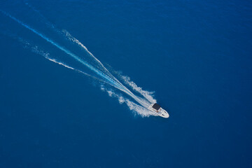 Naklejka na ściany i meble Top view of a white boat sailing in the blue sea. Aerial view of a boat with an awning in motion on blue water. Drone view of a boat sailing at high speed.