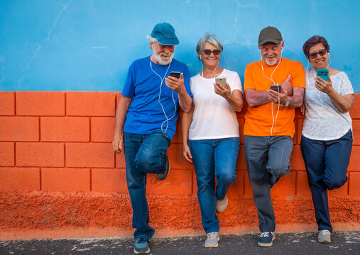 Two Couples Of Senior Brothers And Wifes Looking At Smart Phone Smiling Against A Colored Wall - Four Happy People Using Tech And Social - Active Retirement Concept