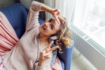 Fototapeta premium Wonderful woman with curly short hairs listen favorite music and lying with closed eyes with pleasure. Wearing cute pink loungewear.