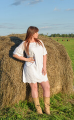 female model in white dress on a field with haystacks. a beautiful young woman with brown hair in short shorts and a white shirt. harvest concept