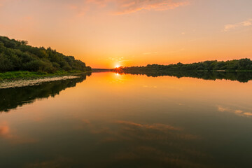 Fototapeta na wymiar Scenic view at beautiful summer river sunset with reflection on water with trees , golden sun rays, calm water ,deep blue cloudy sky and glow on a background, spring evening landscape