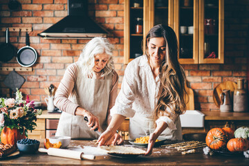 Young woman and mother in white aprons stand at large brown wooden kitchen table and prepare tasty...