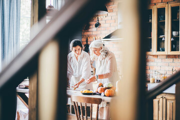 Young woman and mother in white aprons stand at large brown wooden kitchen table and prepare tasty dinner. Senior mother and happy adult daughter baking pumpkin pie together at home.