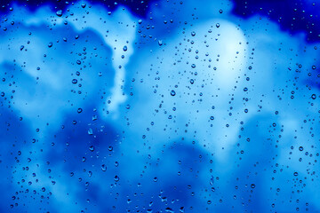 Naklejka na ściany i meble blue rain water drops on a window glass close up with blue colorful clouds in background, drop and cloud macro texture ; bad weather backdrop
