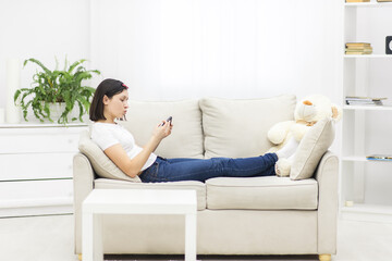 Photo of beautiful mother lying on sofa in living room and holding phone.