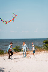 Kid holding kite near golden retriever and parents playing football on beach