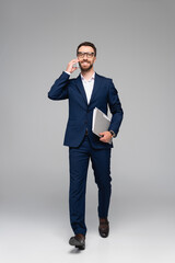 full length view of businessman in blue suit talking on smartphone and holding folder while walking on grey