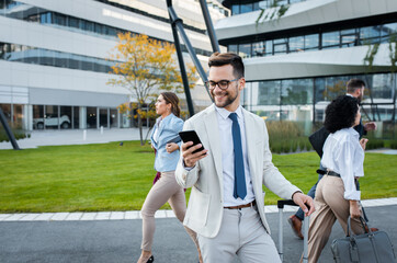 Portrait of smiling businesswoman with smartphone in his hands standing in front of modern office buildings with colleagues behind him.