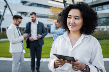 Fototapeta premium Portrait of smiling asian businesswoman with tablet standing in front of modern office buildings with colleagues behind her.