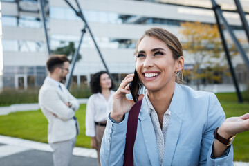 Portrait of smiling businesswoman talking at smartphone while standing in front of modern office buildings with colleagues behind her.