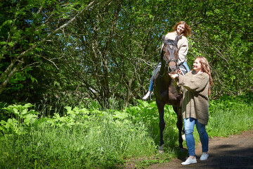 Fat and slim girls walking with brown horse in a Park on a Sunny summer day and green trees in the background. Two young female friends or sisters riding and having fun and joy with a horse on nature.