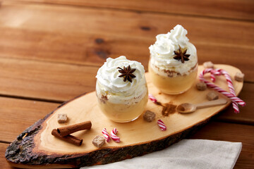 christmas and seasonal drinks concept - glasses of eggnog with whipped cream topping, candy canes, sugar and cinnamon on wood cut board