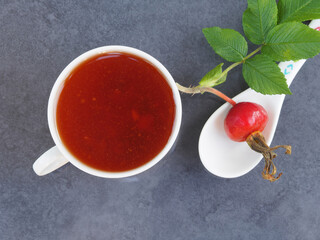 Healthy drink of wild rose in a white cup and saucer with the berries in white spoon on a dark table, top view. Delicious tincture of seasonal fruits with vitamins. Herbal tea with rosehip