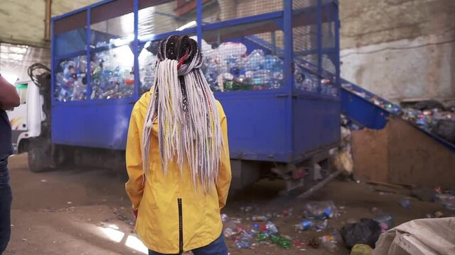 Backside footage of a woman with dreadlocks wearing yellow jacket watching the process on waste, recycling factory. Big truck with cage carcase of used plastic bottles for recycling