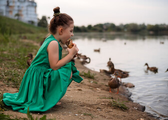 Little girl 5 years old in a beautiful green dress feeds the ducks with bread on the river bank