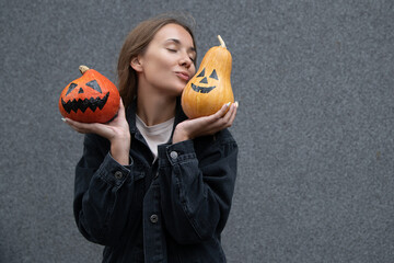 Young attractive European female wearing dark jacket, holding and kissing pumpkin on gray...