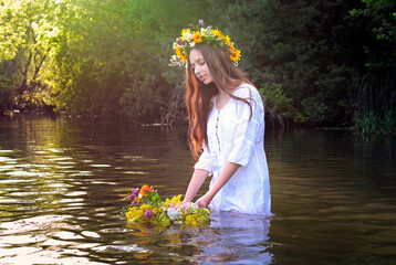 A young slim girl European appearance in a white shirt stands in the river and holds a wreath of their yellow flowers