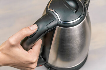 Obraz premium woman's hand holds the black handle of a gray electric kettle and presses the power button close-up, top view