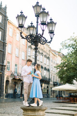 Fototapeta premium Romantic date in old ancient city outdoors. Happy young stylish couple in love with cups of take away coffee, standing under the street light and looking at each other