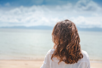 A woman with curly hair stands with her back in the wind on the beach. Sea and travel, hair care