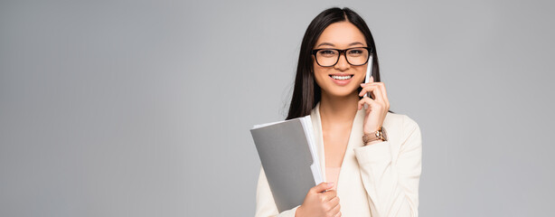 horizontal image of asian businesswoman in eyeglasses holding folder and talking on mobile phone isolated on grey
