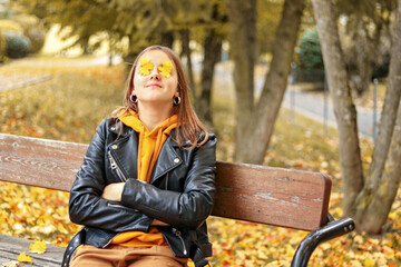Young teenager girl relaxing and meditating in autumn park sitting on wooden bench with yellow maple leaves on her eyes. In love with fall.