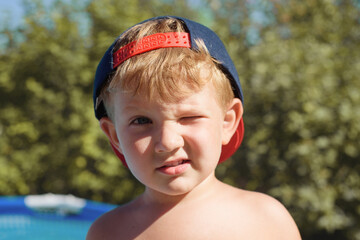A little boy in a cap squints from the sun and looks at the camera. Close-up - obrazy, fototapety, plakaty