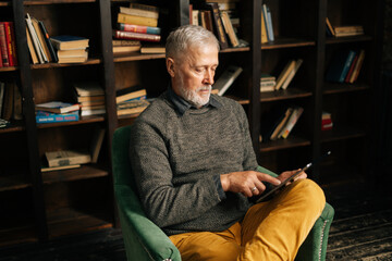 Focused bearded gray-haired mature adult male using digital tablet , sitting at home on background...