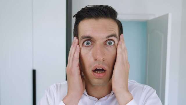 Scared businessman looks into camera with startle at face clutches his head showing fear and surprise. Portrait of shocked man on office background.