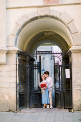 Fototapeta na wymiar City outdoor portrait of happy young couple in love, posing near pod building with beautiful vintage gate. Pretty girl in blue dress holds bouquet of red flowers, handsome guy kisses her